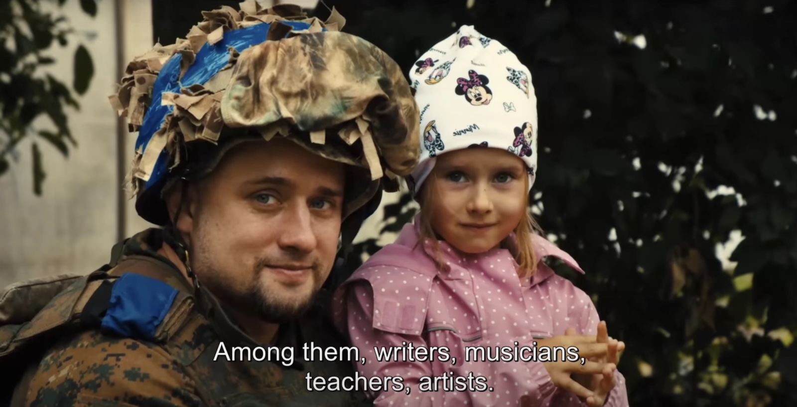 A soldier poses next to a child with blond hair and a pink jacket. The subtitles across the screen read, 'Among them writers, musicians, teachers, artists.'