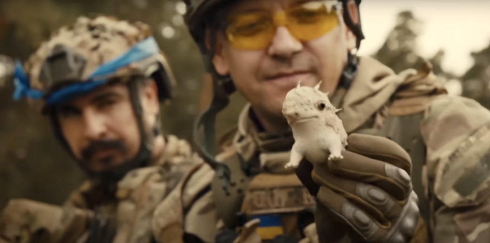 Two men in military fatigues and helmets look towards a small white porcelain creature held in the gloved hands of the man on the right. The whimsical creature has big eyes and spikes.