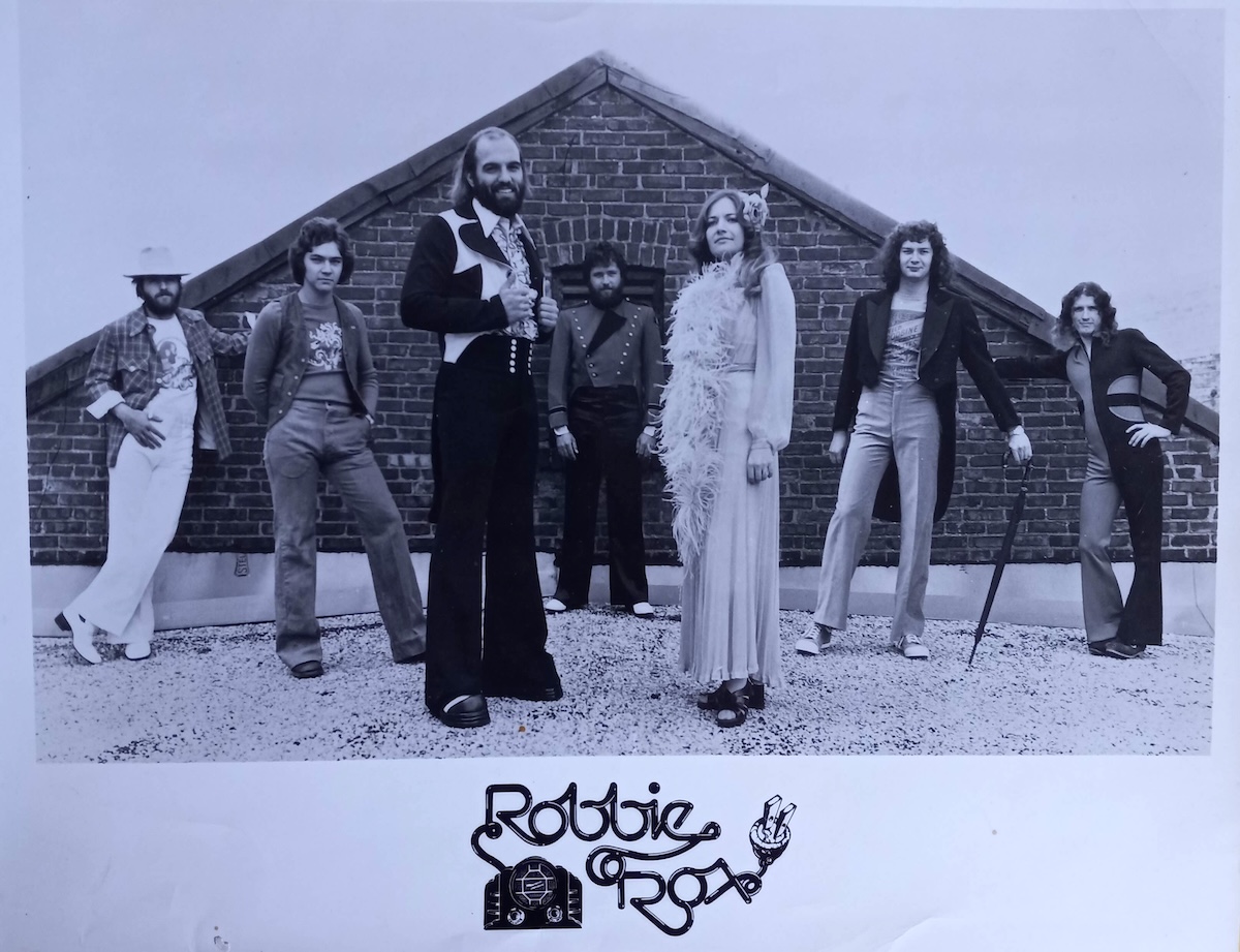 A group of seven musicians poses together in front of a triangular brick structure. They have long, wavy hair and are attired in the colourful suits, feather boas and flared trousers of the early 1970s. The black and white photo is set against a light blue background.