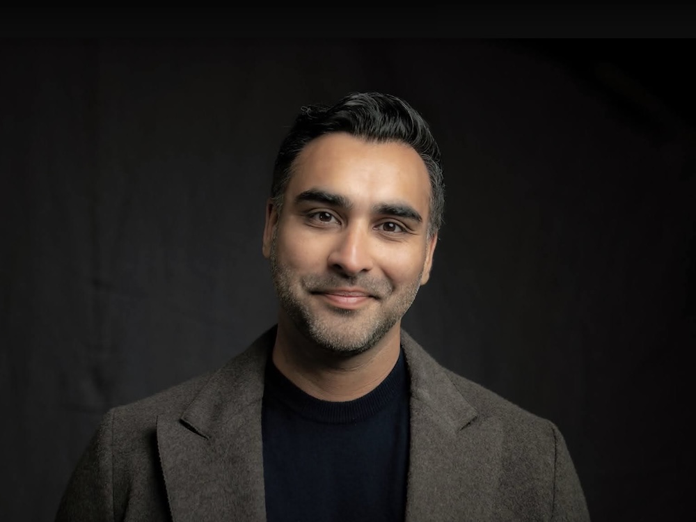 A man with medium skin tone and short black hair and a salt and pepper beard wears a black shirt and a grey-brown felted suit jacket. He is smiling at the camera.