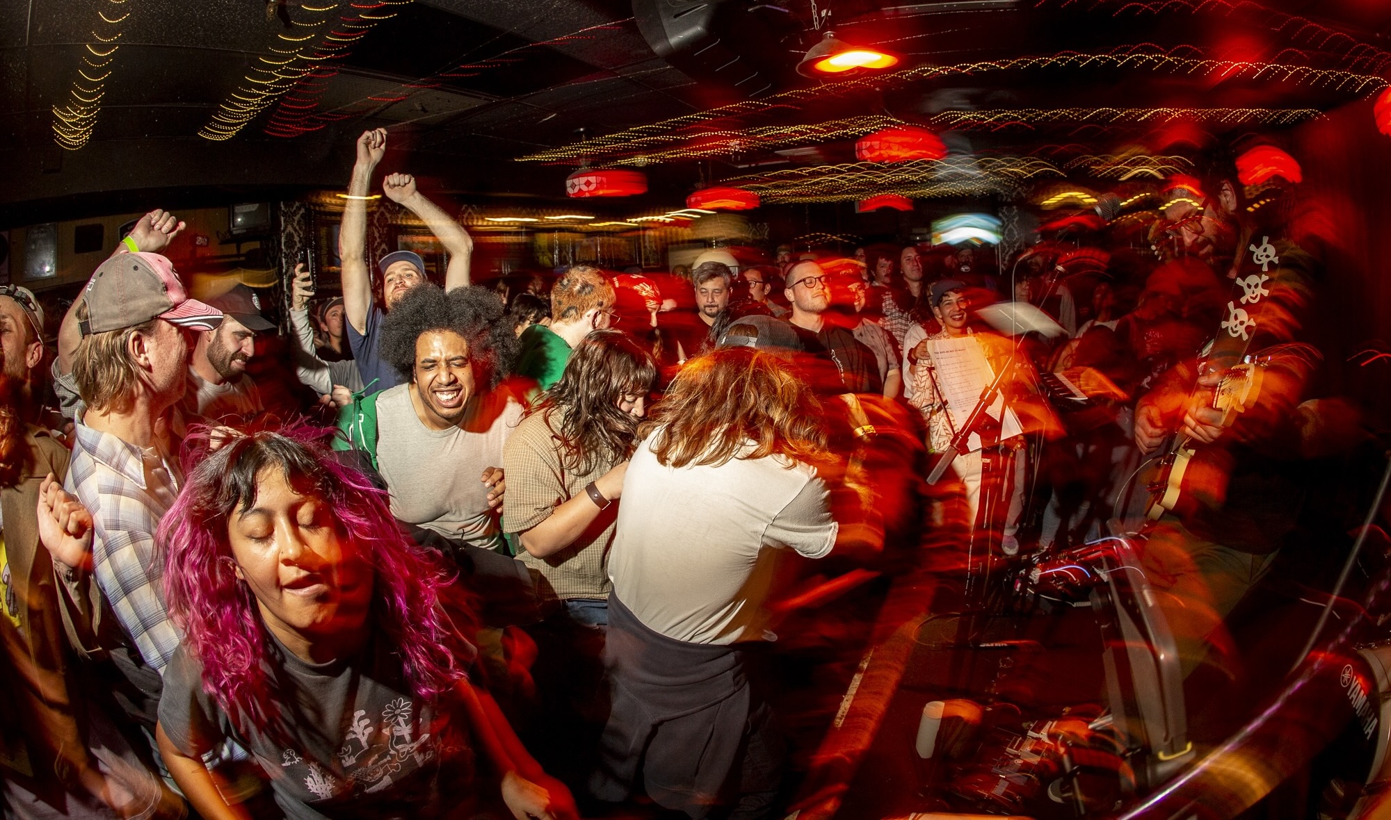 A densely packed indoor space features streaks of red and yellow light. On the right, a band is playing on a small stage; they are abstracted and distorted using camera effects. On the left, the crowd is moving to the music; a diverse group is attired in casual streetwear, moving closely and joyfully together.