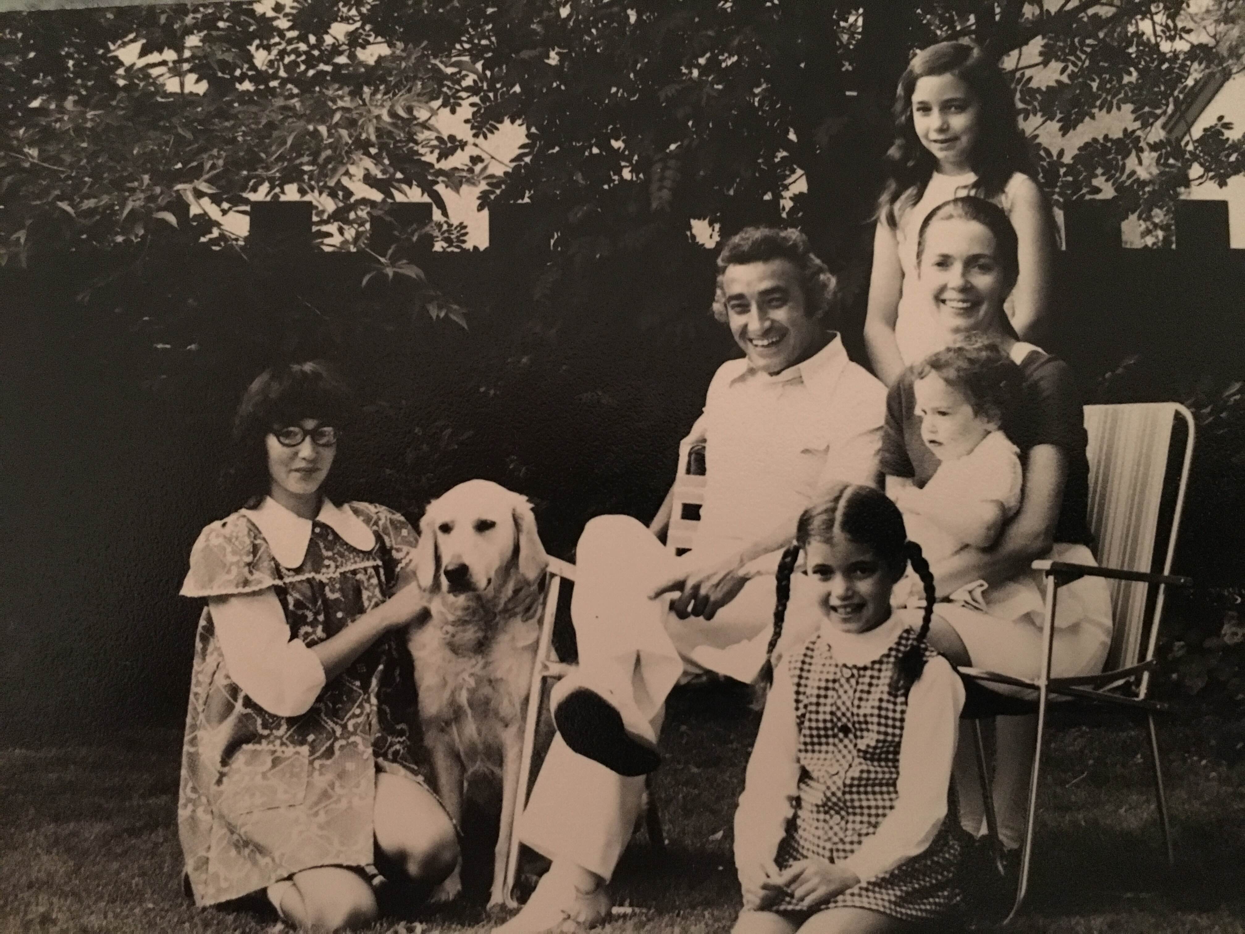 A black and white photograph from 1972 features Mel Hurtig seated on a lawn chair in a leafy yard. He is wearing white and smiling brightly. Around him are his family members: his spouse is seated beside him, holding a toddler in her lap. Three older girls are seated in front of, behind and beside their parents, the daughter on the left with the family dog.