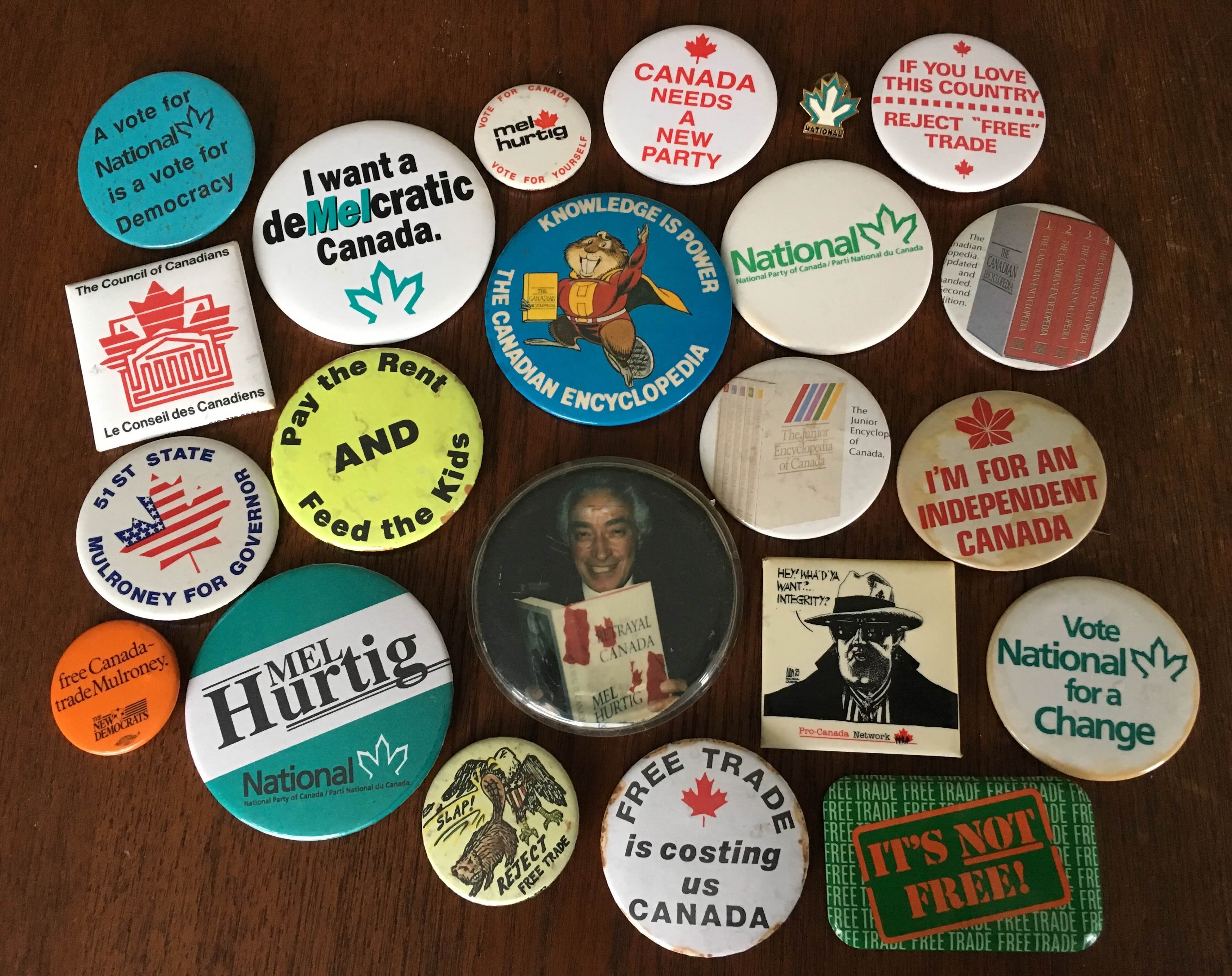 A colourful collection of round buttons are laid out on a wooden tabletop. They include political campaign buttons for Mel Hurtig alongside buttons that advocate for Canadian sovereignty, positive social change and the rejection of free trade.