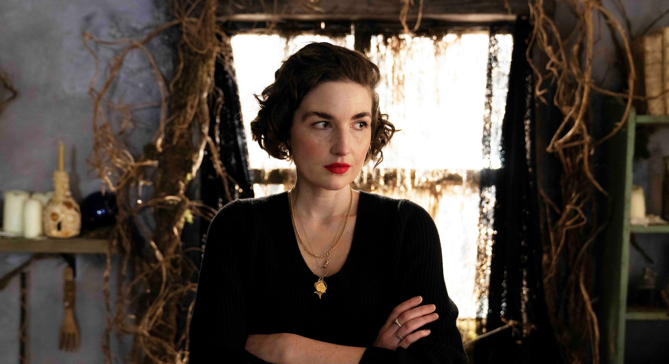 Elizabeth Sankey has short brown wavy hair tied back in a partial updo. She is wearing several gold necklaces and a black top. Her arms are folded and she is looking to the side, towards the right of the screen. Behind her is a dense indoor space that resembles a root cellar: a window shining bright light is behind Sankey, and tangles of roots cascade down each side. The walls are painted blue and a shelf of candles and vases lines the wall on the left.