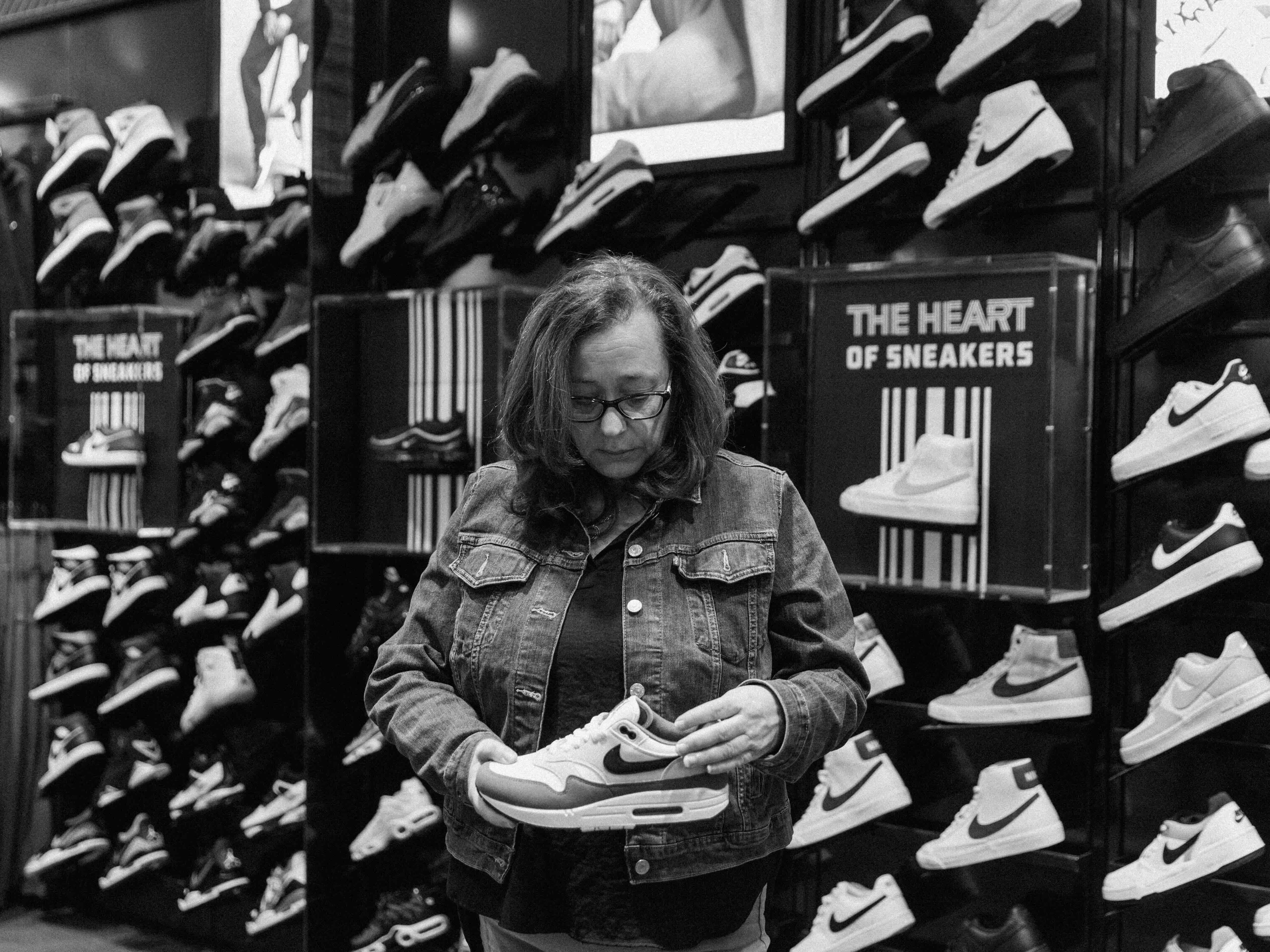 Traci Letts stands in front of a wall displaying dense, angular rows of sneakers in a retail space. She has wavy shoulder-length hair, glasses and is wearing a denim jacket over a dark top. She looks down while she holds one athletic shoe.