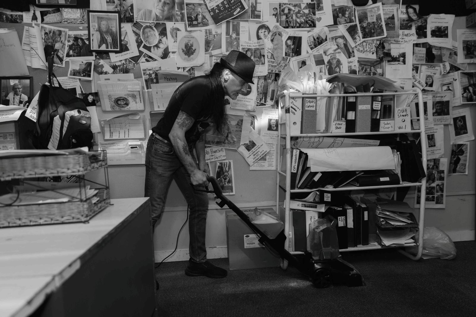 Martin Steward is wearing a fedora over long dark hair tied back. He is wearing a dark T-shirt and jeans and is operating a steam cleaner on the floor of a crowded office space. To his right is a low shelf full of thick binders. Behind him, the wall is covered with printouts of memorial photographs.