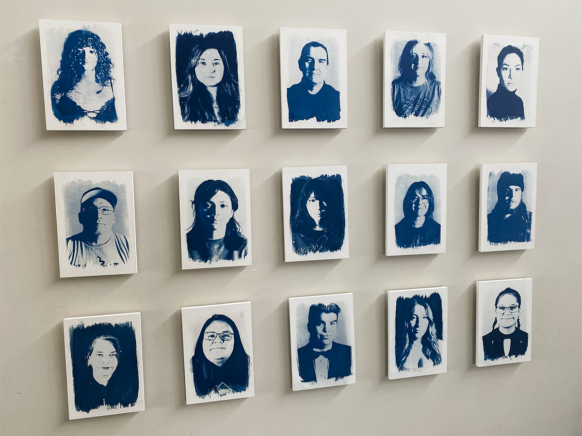 Three rows of blue cyanotype photographs are arranged on a white gallery wall. They are portraits of Vancouver Island University students who are former youth in care.