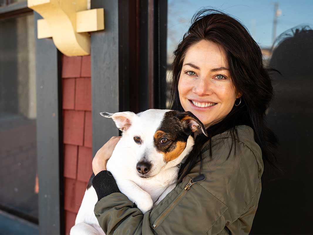 Tarah Dowling has long dark hair and green eyes. She wears an olive-green jacket and stands outdoors against a window with a shingled red wall and black trim. She is looking at the camera and smiling. She holds a small white dog with black and brown markings.