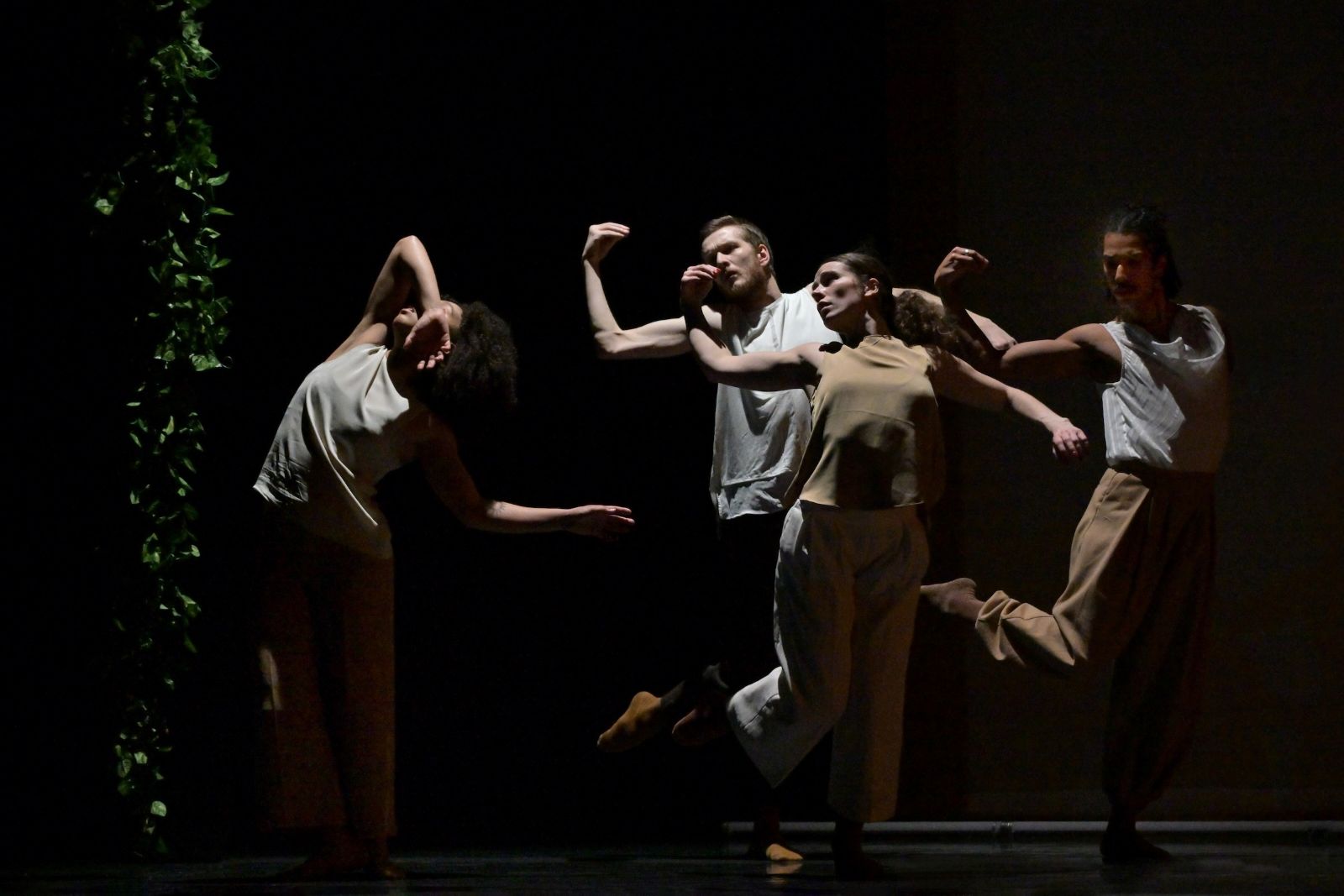 Four dancers move in formation across a stage with a black background. To the left is a column of artificial green leaves. The dancers are clad in loose-fitting sleeveless tops and loose-fitting pants in light neutral tones.