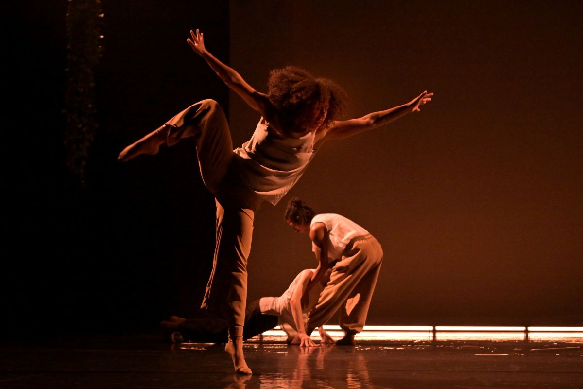 Marisa Gold has dark curly hair. She is a dancer standing on one foot, one leg in the air. Behind her are two other dancers, one lifting the other by the shoulders, on a stage with an orange background.