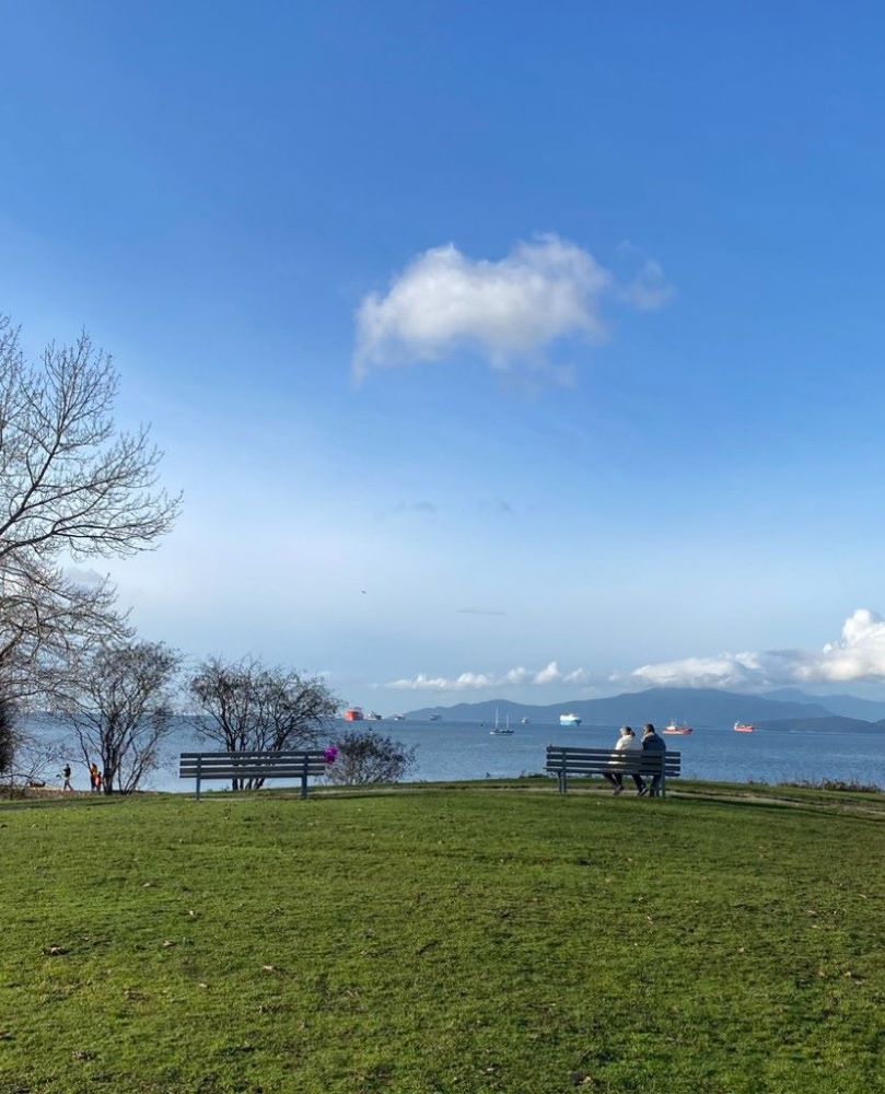 An image of an outdoor view of two benches on a grassy hill against a background horizon filled with the ocean, ships, mountains and a blue sky.