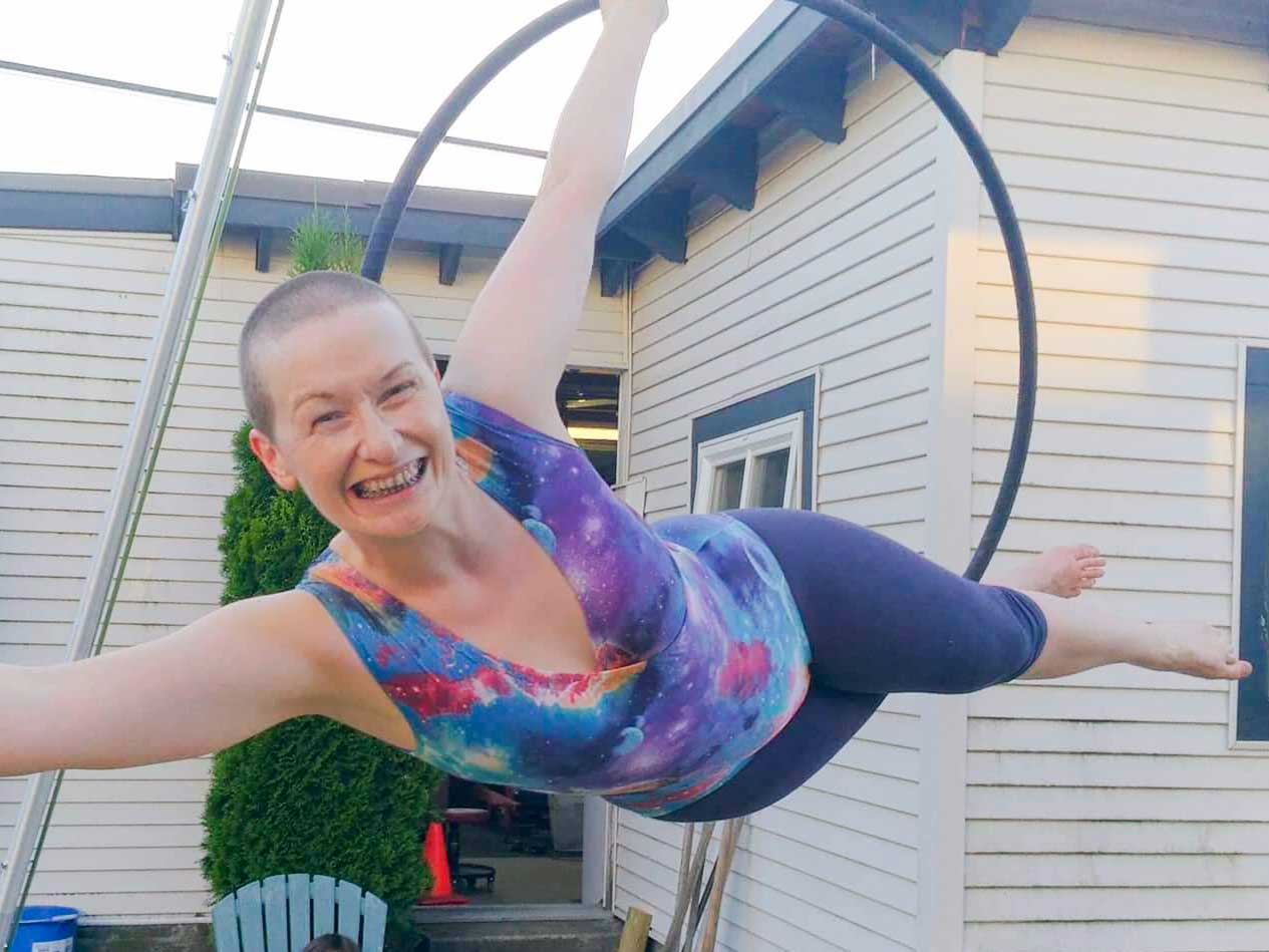 A light-skinned person with a shaved head, wearing a blue and purple leotard, balances horizontally on their side in a suspended steel hoop. 