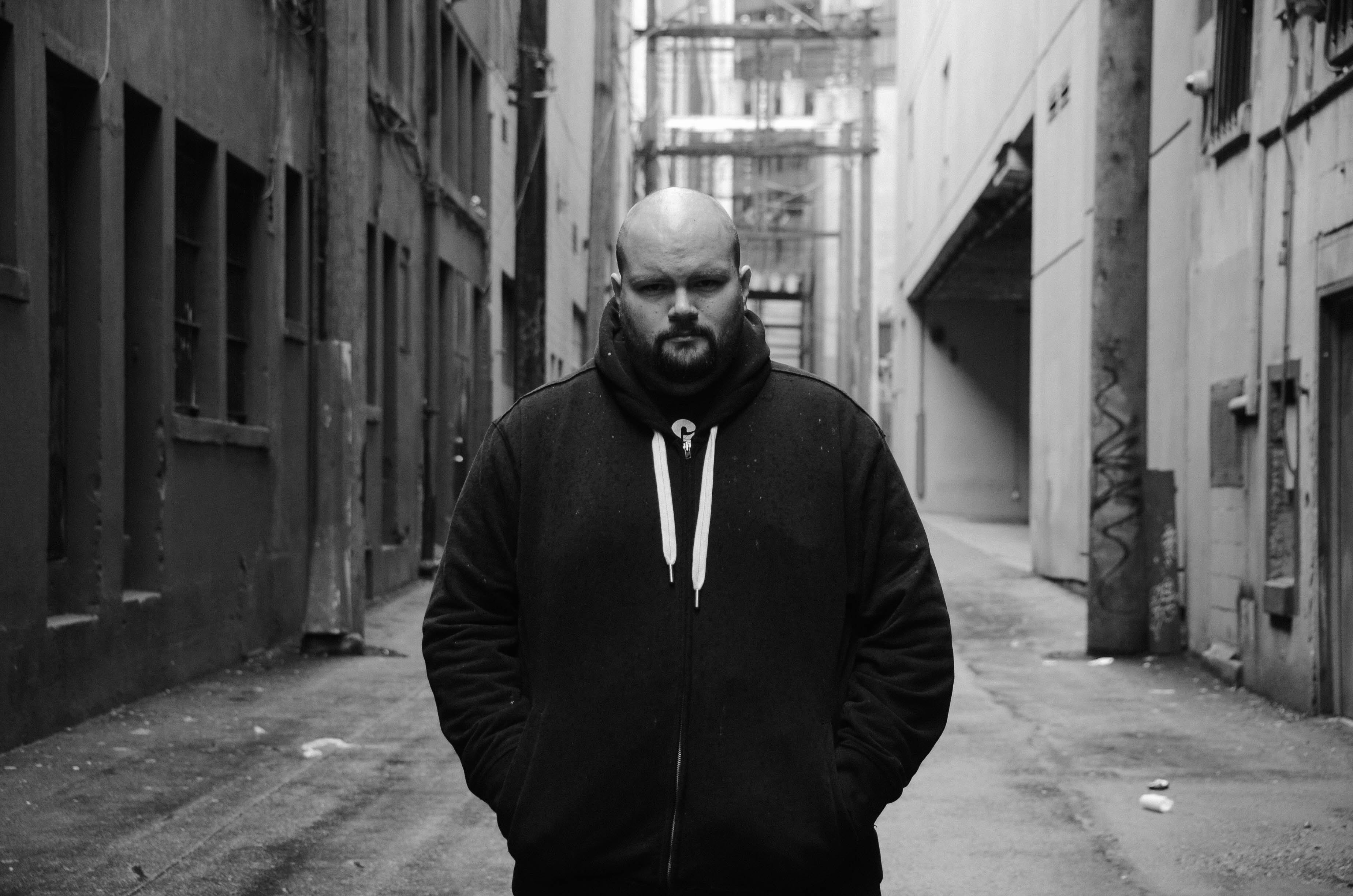 Sam Wiebe stands in the middle of a downtown alley. The black-and-white photograph depicts Wiebe in the centre of the frame, looking directly at the camera. He is wearing a dark hooded jacket. He has a shaved head, light skin and a beard.