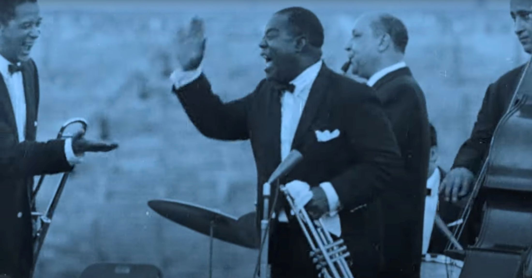 Louis Armstrong is a Black man in a tuxedo. He is holding a trumpet and standing on a stage among other similarly attired musicians. He is smiling widely and making a gesture towards shaking the hand of a man to the left of the frame. The black and white photograph is tinted blue.