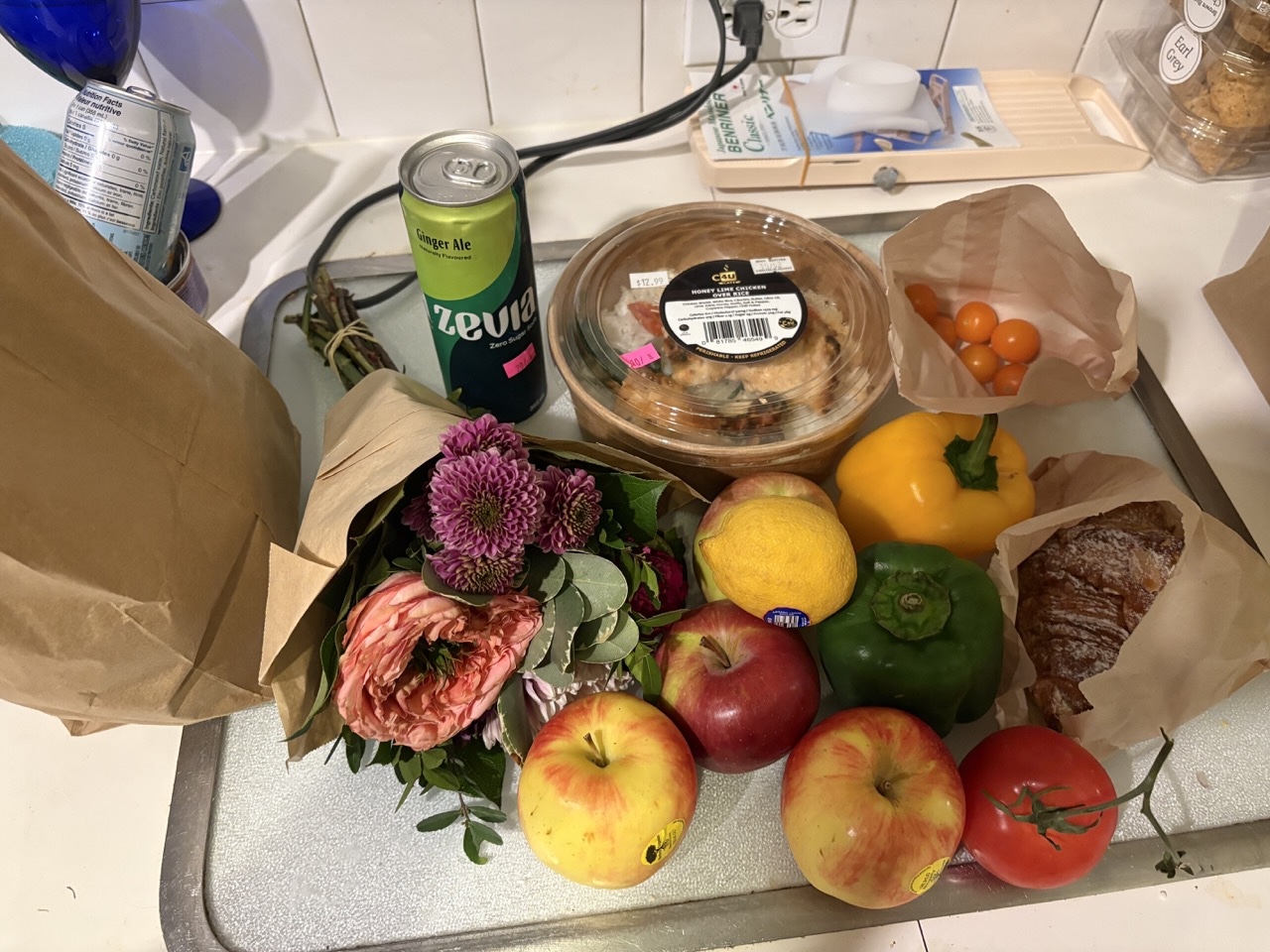 An assembly of grocery items is shot on a kitchen counter from above. To the left, a bouquet of flowers sits next to an assortment of apples, peppers, a croissant and a pre-made meal.