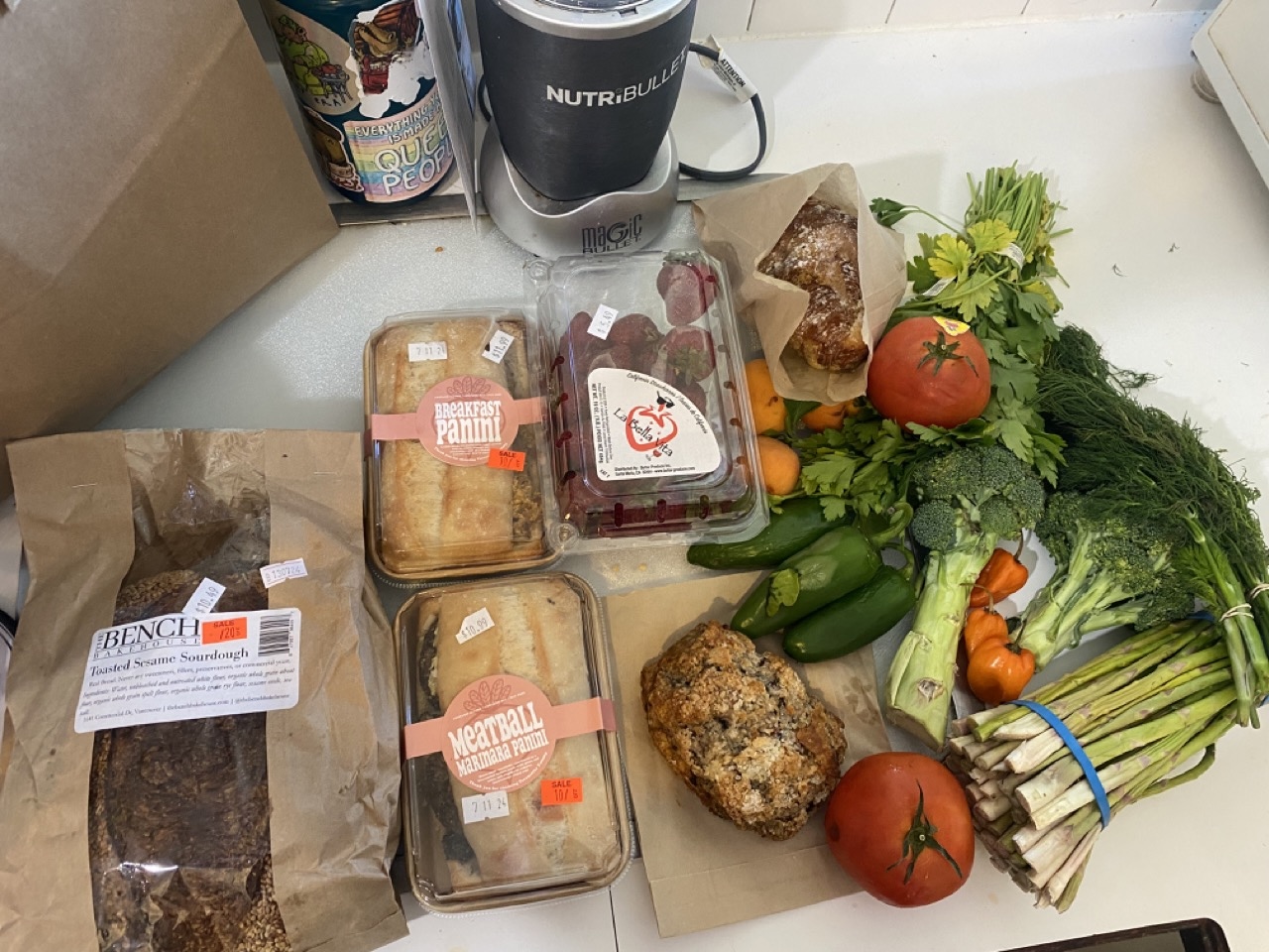 An assortment of grocery items sits on a kitchen counter. They include a packaged sourdough loaf, panini sandwiches and an assortment of produce.