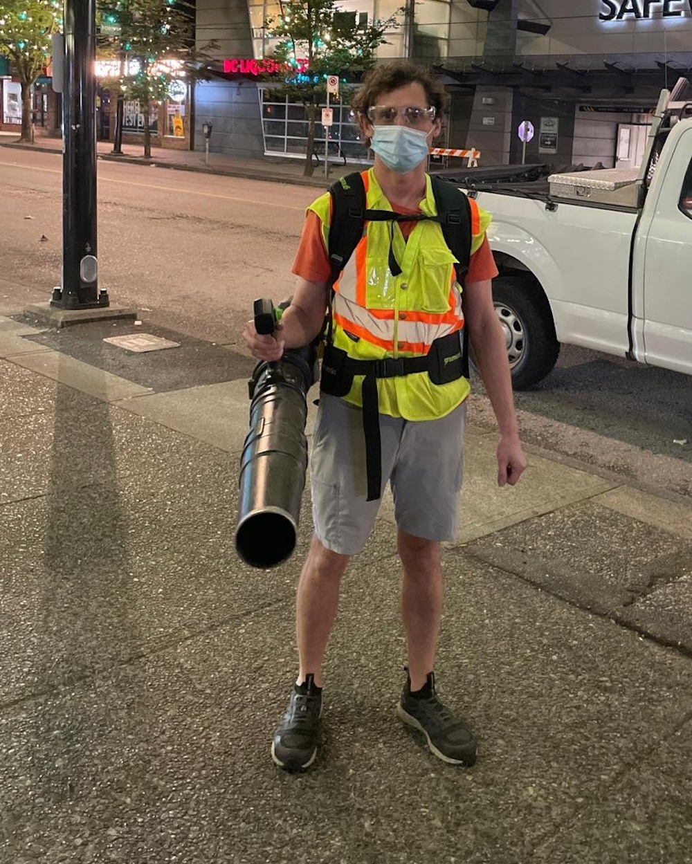 A sanitation worker in grey shorts and a safety vest, wearing a mask and holding a leaf blower.