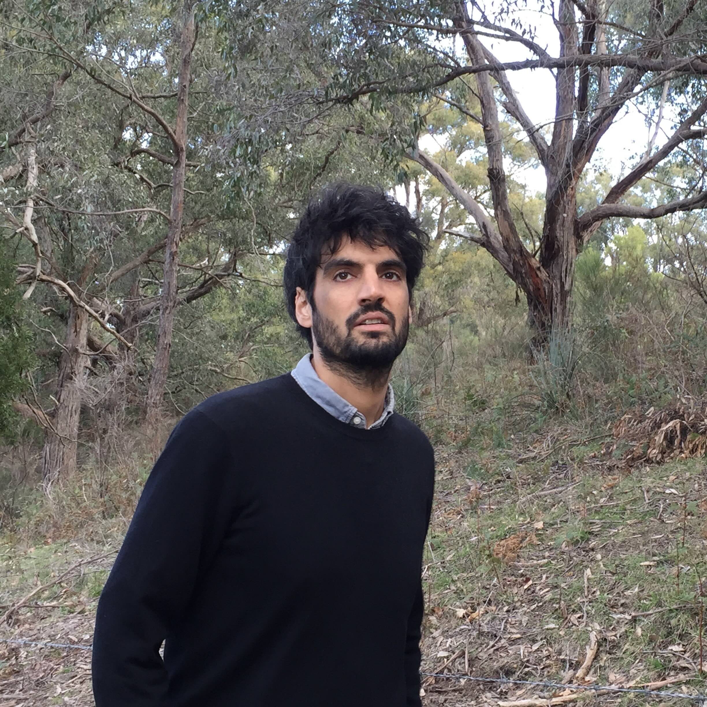 Vijay Khurana has short dark wavy hair and is wearing a light blue button-down shirt under a darker blue sweater. He is standing in a forested setting, looking up and into the middle distance.
