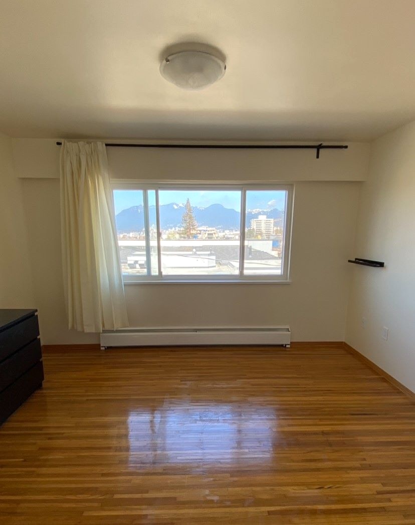 The living room of a bare apartment with hardwood flooring and a view of an industrial-looking roof through the window with the North Shore mountains in the distance.