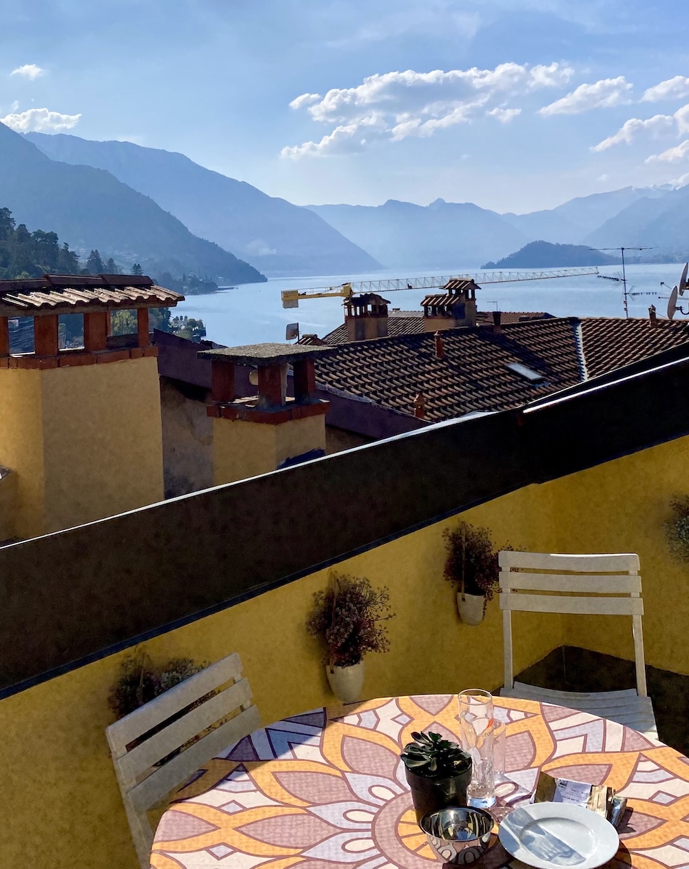 A sunny terrace with a table and chairs, and behind it a view of a lake surrounded by hills.