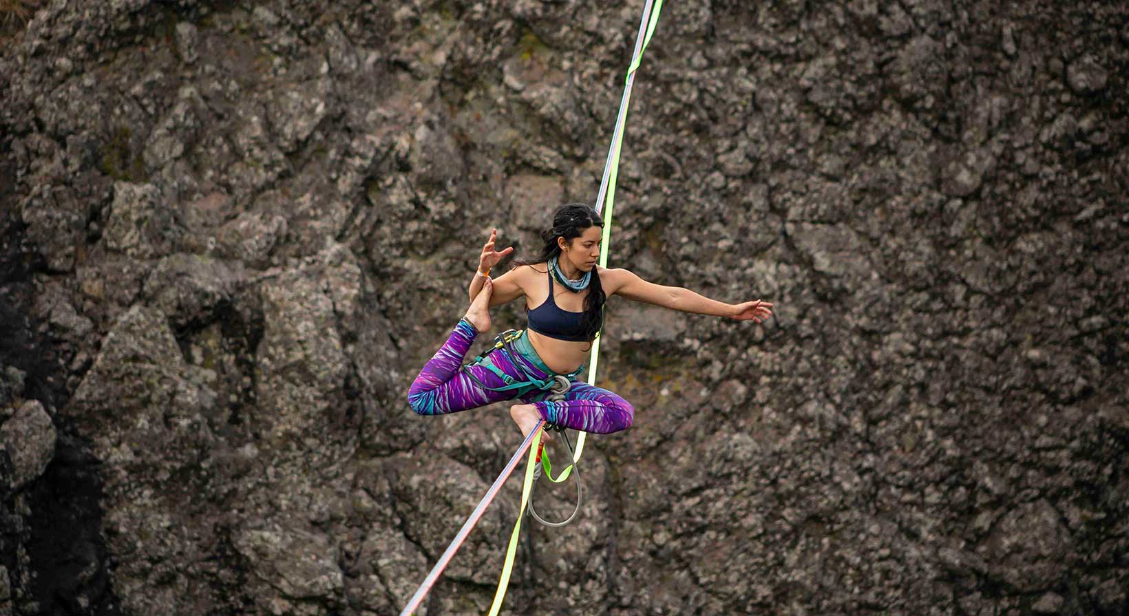 A person with medium-light skin tone and black hair in a ponytail, pulls their leg behind while balancing on a slackline high above the ground.