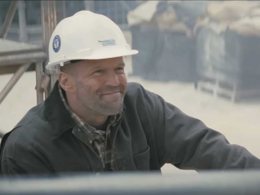 Actor Jason Statham wears a white hard hat and a dark grey work jacket over a muted grey and white plaid shirt. He has pale skin and visible stubble. He is seated in a construction worksite and looking up, smiling.