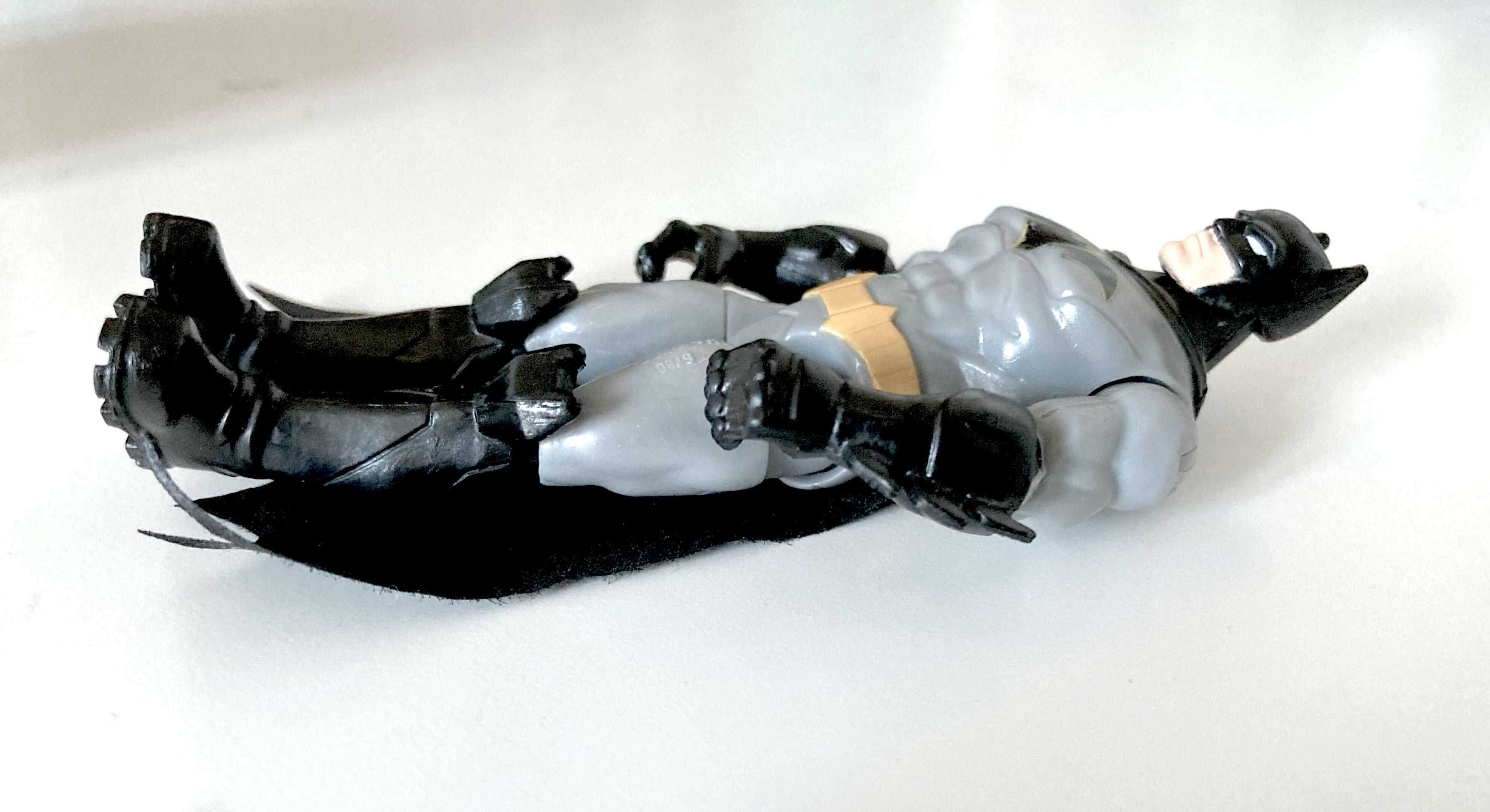 A Batman figurine in a grey suit lies on its back against a white surface. It has a black hood, black cape, gloves and boots, a black bat logo on its chest and a pale yellow belt.