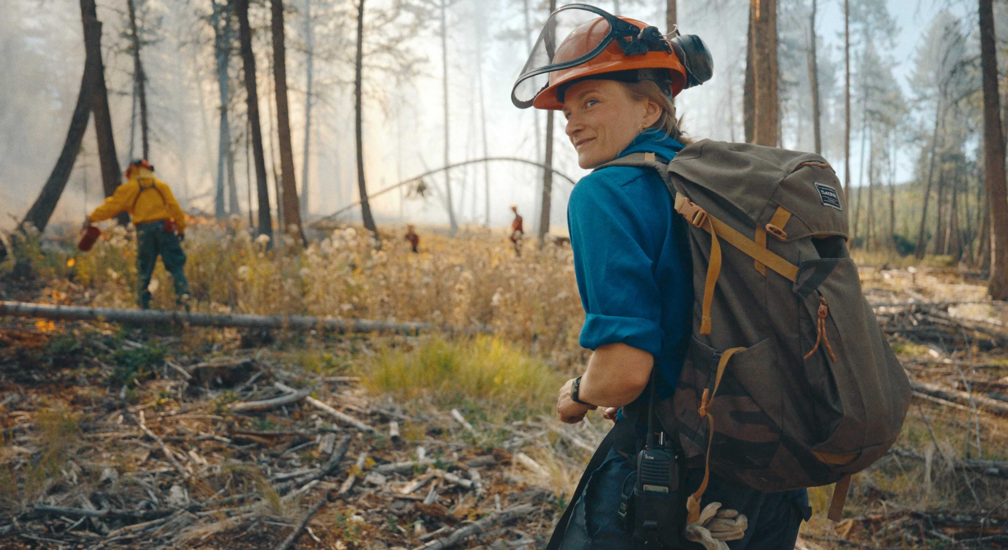 Chloe Kuch is wearing an orange hard hat, a blue button-down shirt and a backpack as she walks through a forest shrouded in smoke. Her back is turned to the camera and she faces the left of the frame, smiling.