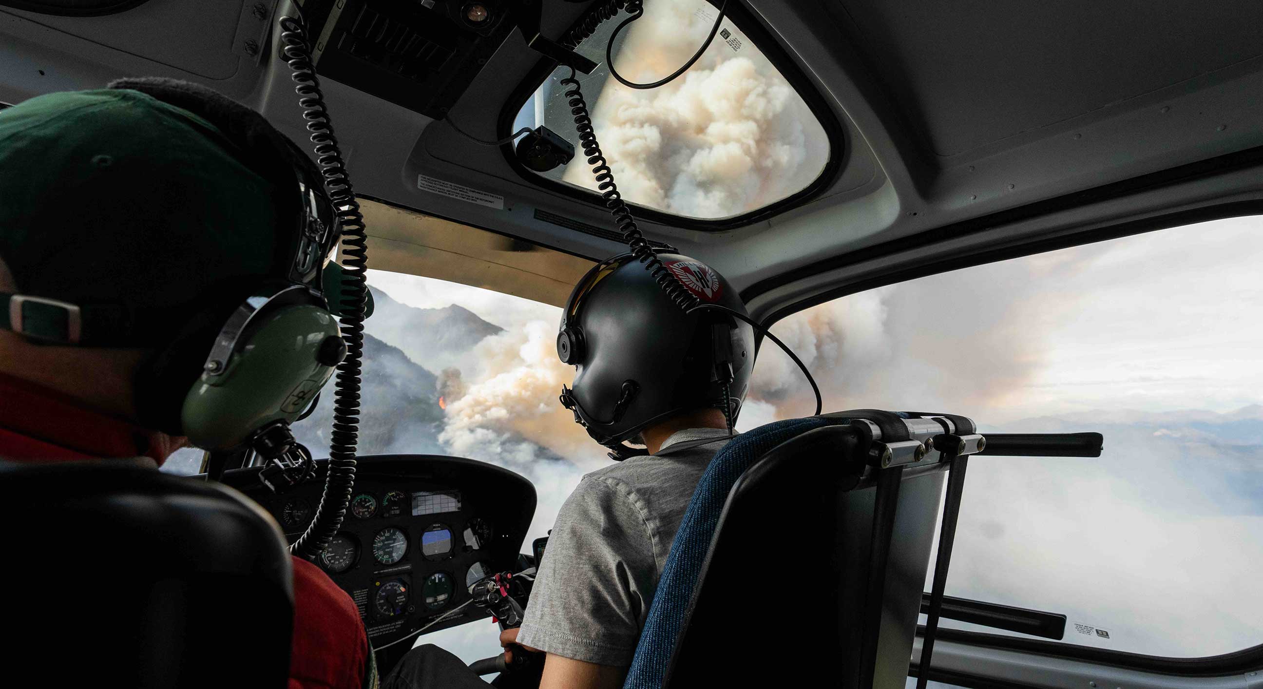 Two people sit in the cabin of a helicopter as it flies over a mountainous area engulfed in thick plumes of white smoke. They are wearing helmets and large headsets, and their backs are turned to the camera.