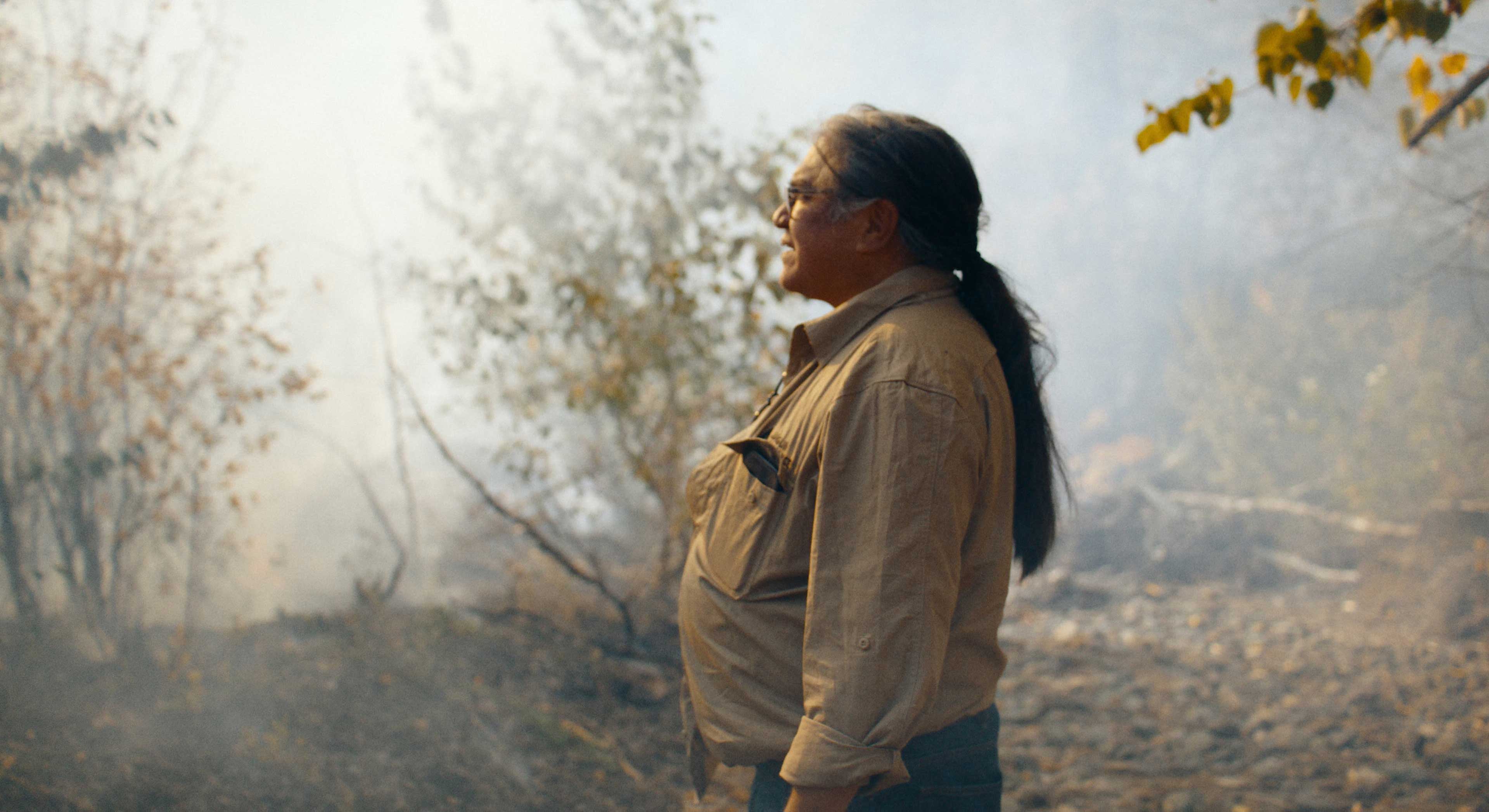 Joe Gilchrist has long dark greying hair pulled back. He is wearing glasses and a tan button-down shirt. He looks to the left of the frame, where the shrubbery around him is shrouded in smoke.
