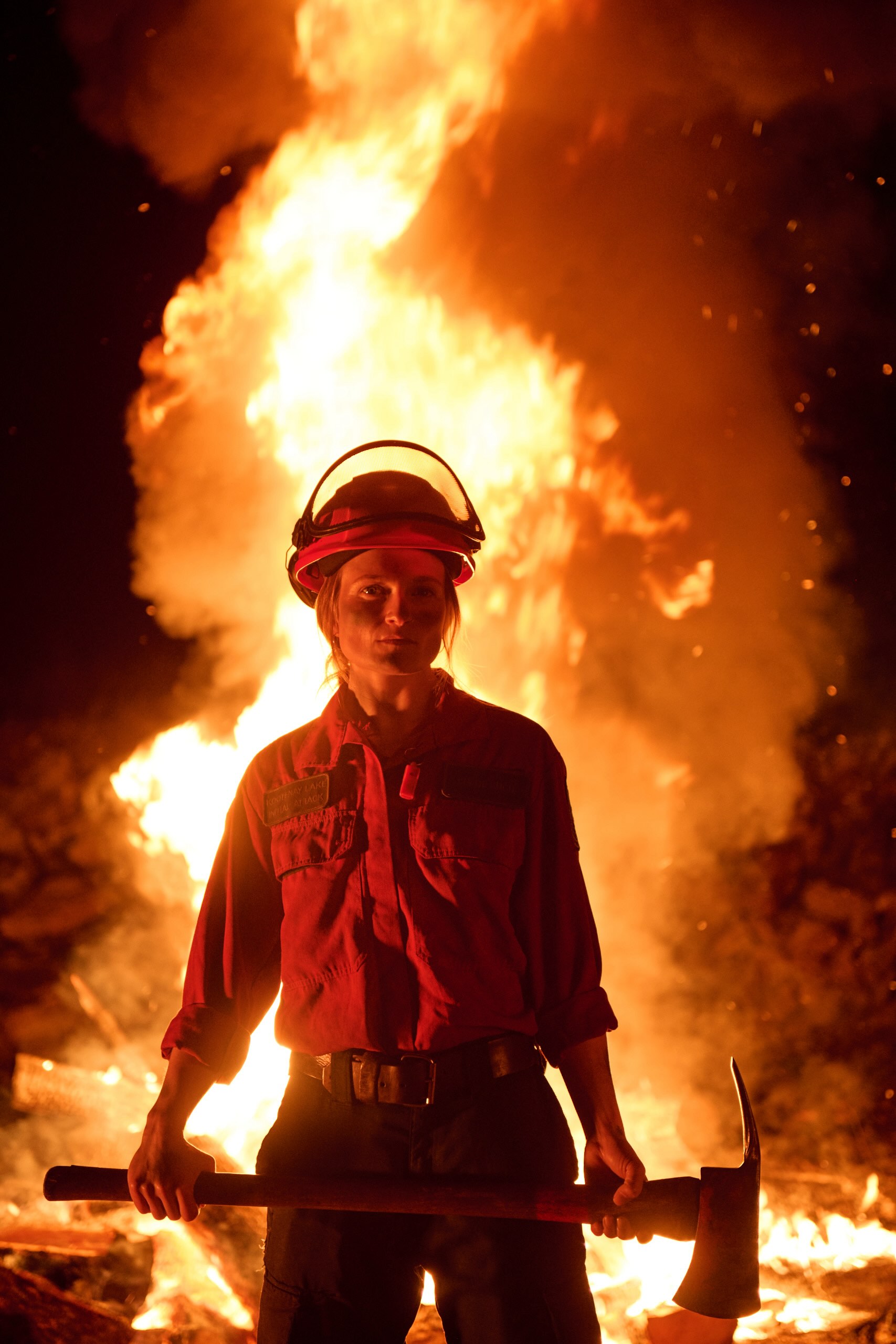 Chloe Kuch is wearing a hardhat, a button down shirt and is holding an axe low across her body. Her light hair is tied back and she is standing in darkness in front of a tall fire.