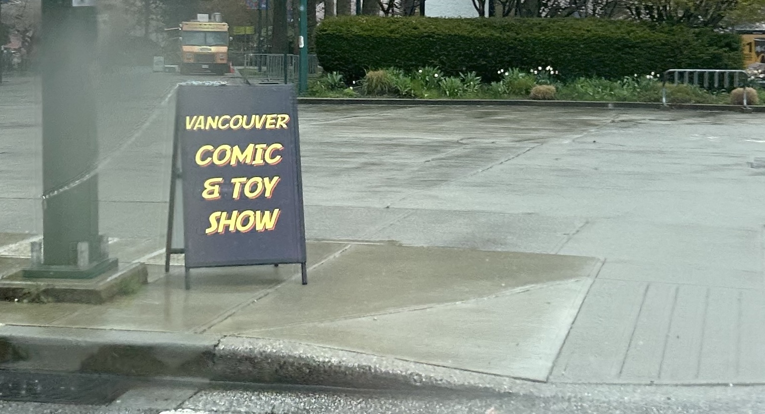 A small black sandwich board sits on a sidewalk on a rainy day. It reads “Vancouver Comic & Toy Show” in yellow text with a red drop shadow.