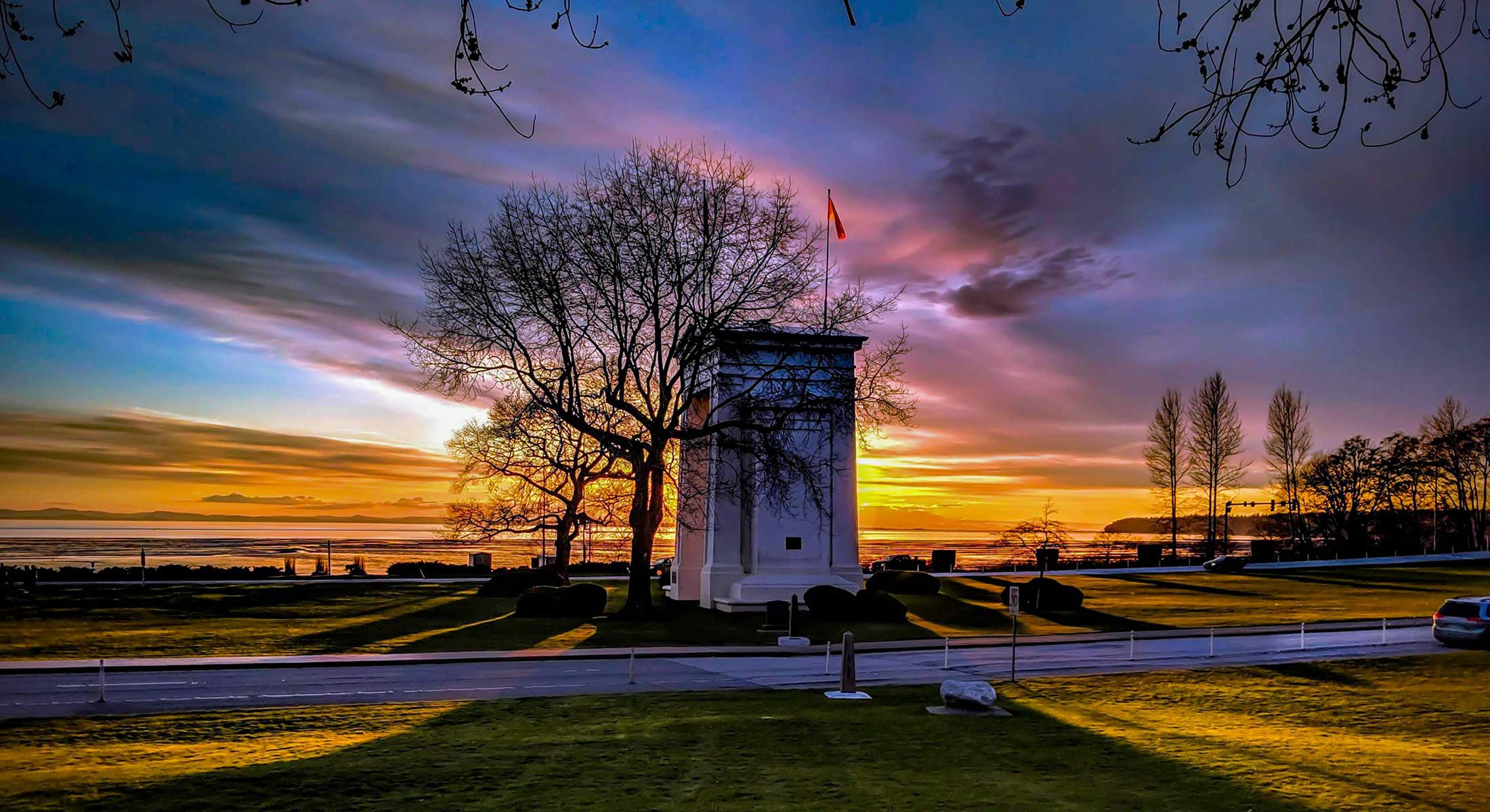 The view of the side of a tall white monument in the middle of a park is obscured by the branches of deciduous trees, which are visible in silhouette against a colourful sky at sunset.