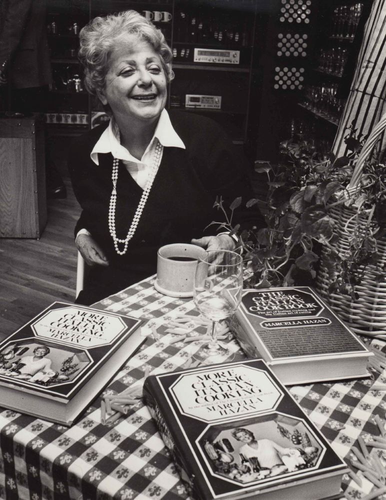 A black-and-white photo of Marcella Hazan finds her seated at a small table with checkered tablecloth, a coffee cup, a wine glass and three copies of her cookbooks.
