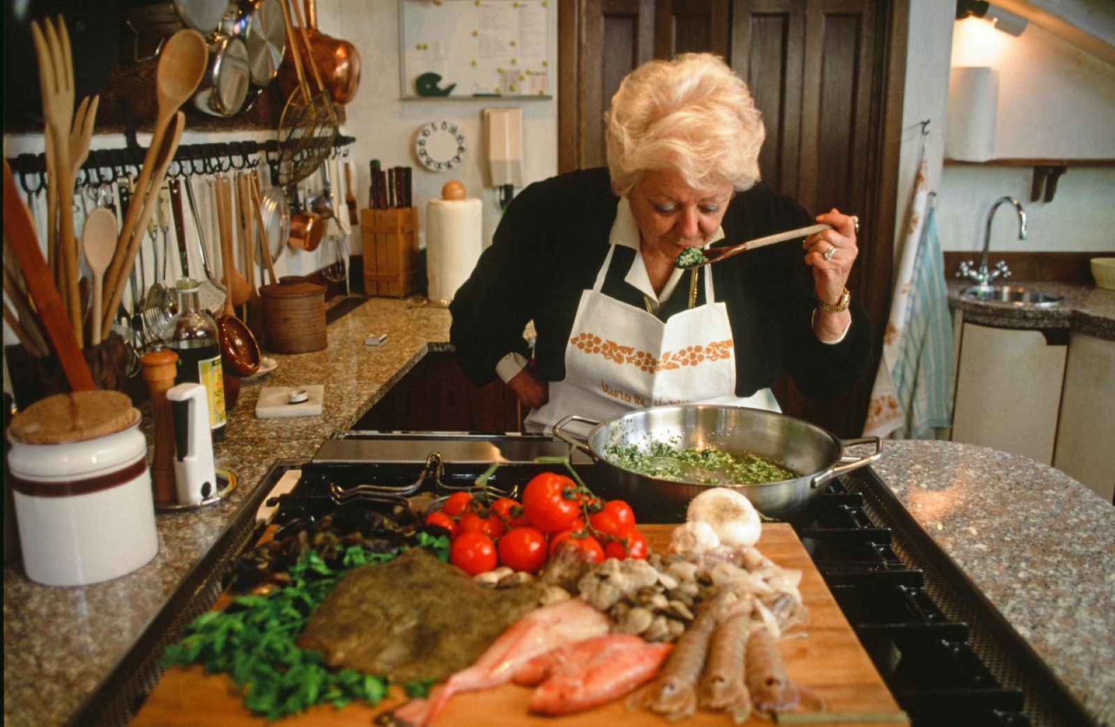 Marcella Hazan is standing in a kitchen. She leans over a silver saucepan to taste the greens on a wooden spoon. In front of her is a wooden cutting board festooned with tomatoes, garlic, greens and seafood.