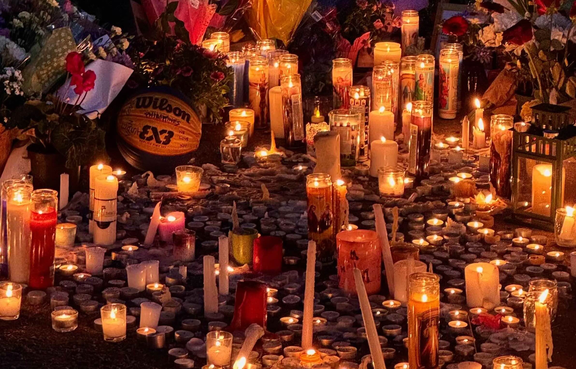 Tea lights, tall white candles and other candles in glass containers are assembled across an outdoor area adorned with bouquets. A basketball with a memorial message sits in the upper left quadrant of the frame.