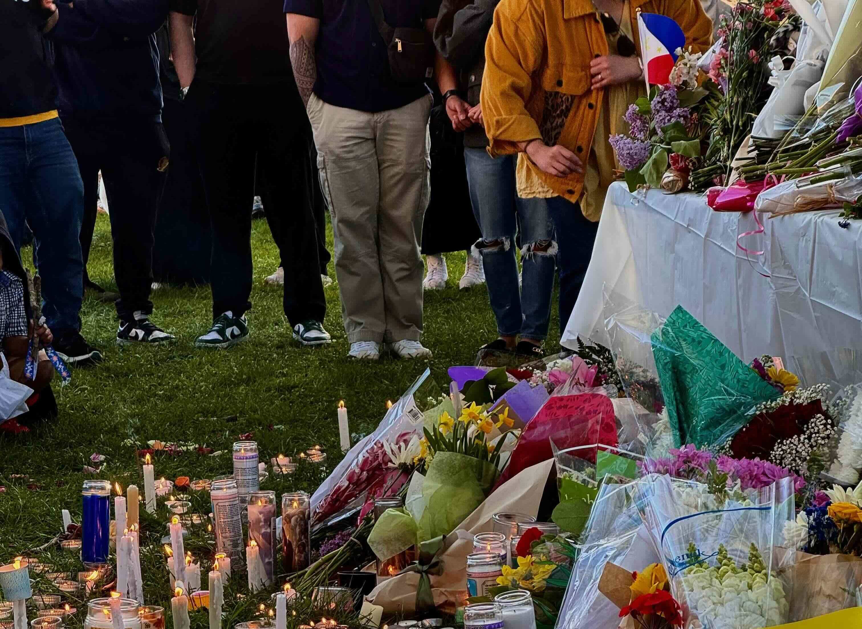 People stand in the grass beside a memorial display consisting of flowers, candles and a table loaded with flowers and the flag of the Philippines. People are visible from their chest down, but we can’t see their faces.
