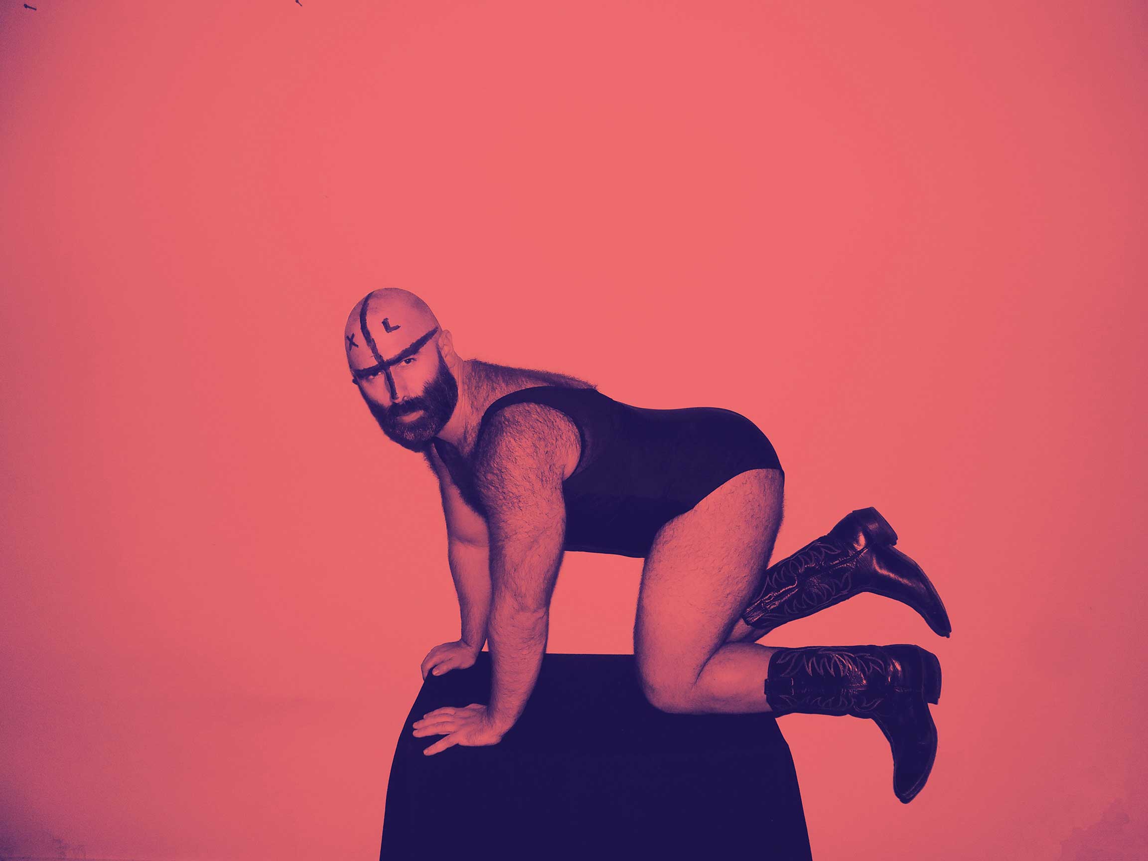Jeff Cancade poses in a black-and-white photograph with a red colour treatment. They are on their hands and knees on a small dark surface wearing greasepaint on their face and head, a short dark bodysuit and cowboy boots.