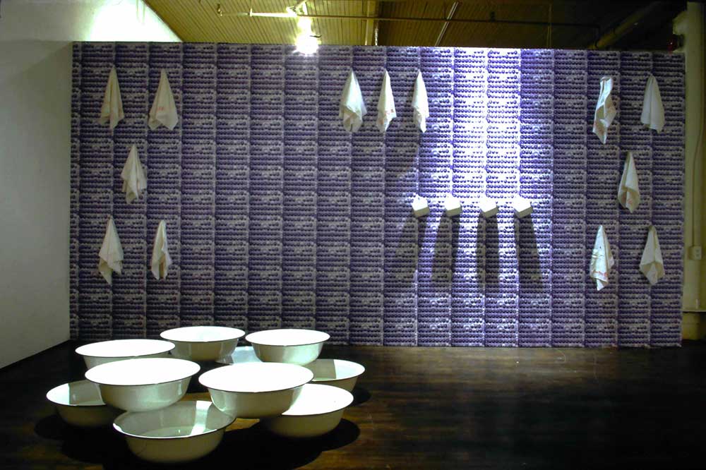 Several large white washbasins are arranged on the lower left quadrant of the frame against a blue and white patterned wall on which white cloths are hung in an indoor gallery space.