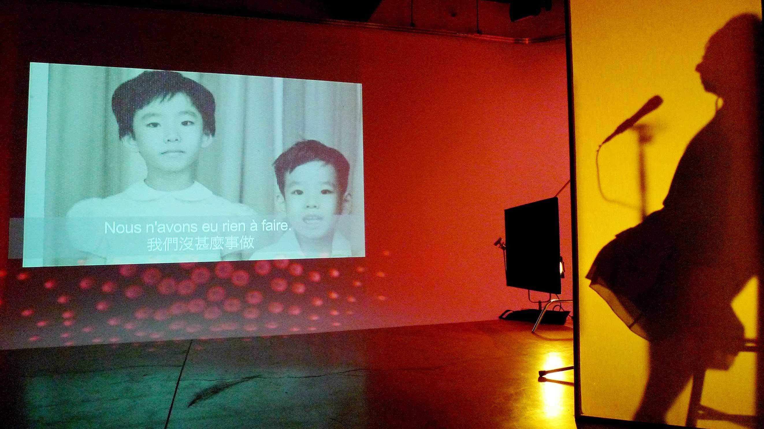 A photograph features a black and white archival image of two Chinese children in a darkened room. To the right of the frame an older man is seated at a microphone and he is seen from the side, in silhouette.