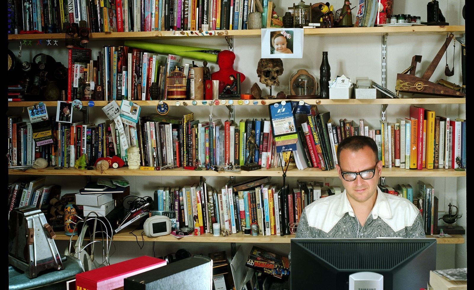 Cory Doctorow sits in front of a computer monitor with a wall of shelves loaded with books, photos and knickknacks behind him.