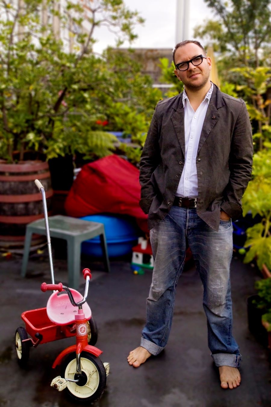Cory Doctorow, a man with light skin tone, close-cropped hair and black-framed glasses, stands barefoot in jeans next to a child’s red tricycle.