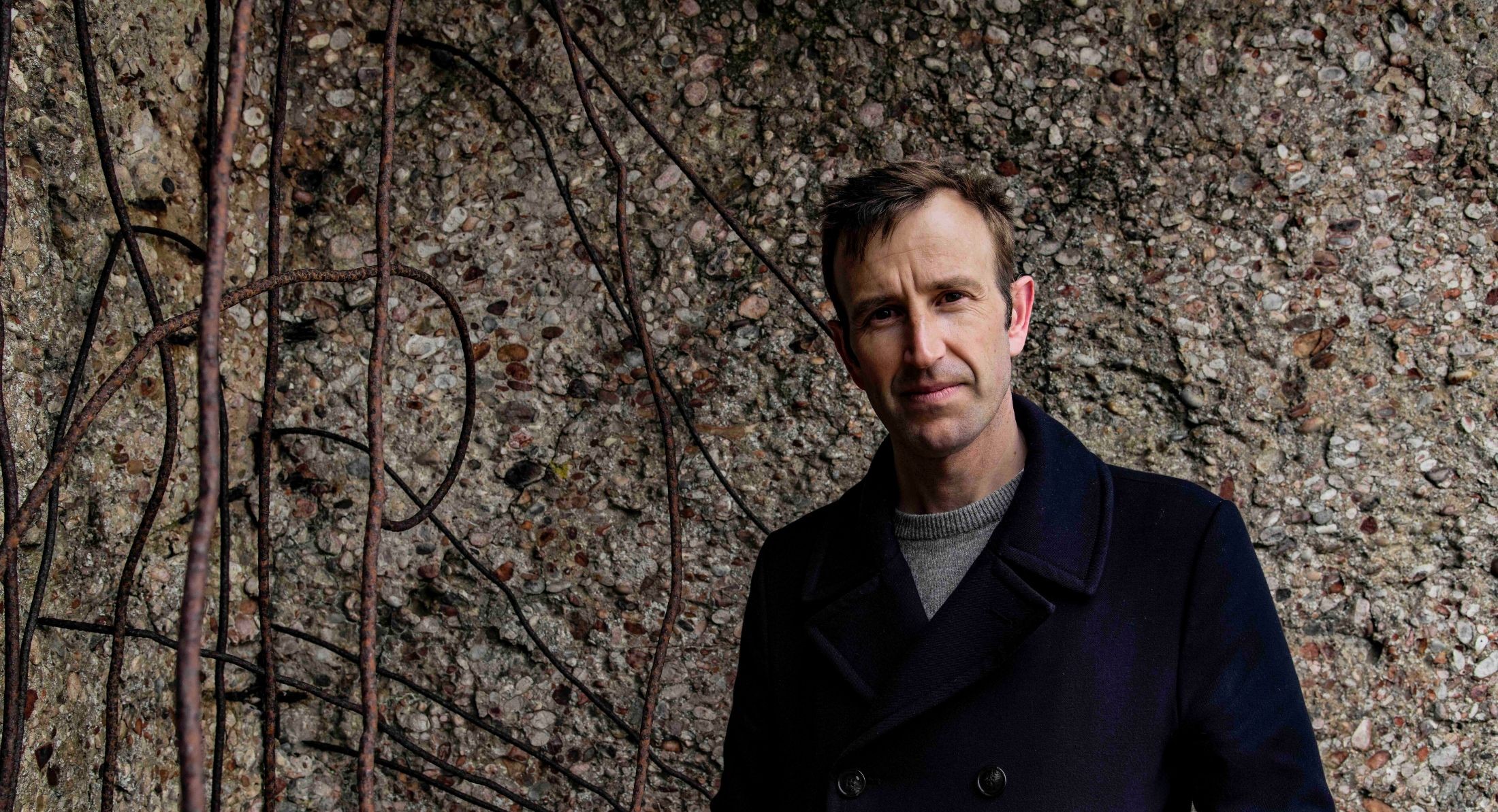 Robert Macfarlane has short brown hair, a pale skin tone and is wearing a dark pea coat over a grey sweater. He is standing against a grey textured wall embedded with stones. Branches cross the background to the left of the frame.