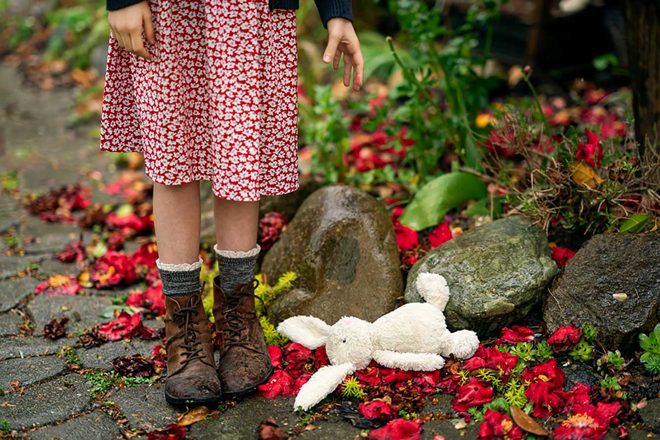 A young girl is photographed from the waist down. She is wearing a red floral dress and has dropped a stuffed white rabbit on the ground. The ground is rocky and lined with red flower petals. There is green foliage in the background.