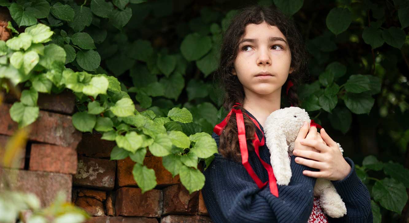 A girl with dark wavy hair tied with a red ribbon holds a white stuffed rabbit to her chest. She has a light skin tone and is wearing a thick navy cardigan over a red floral dress. She is standing against a brick wall with foliage around her.