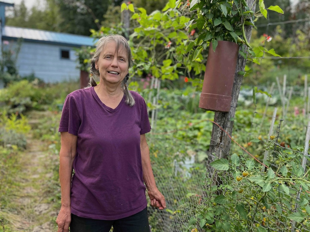 Lois Bradley has greying shoulder-length hair in pigtail braids. She is wearing a purple T-shirt and has a medium skin tone. She is standing outdoors in a garden and smiling.