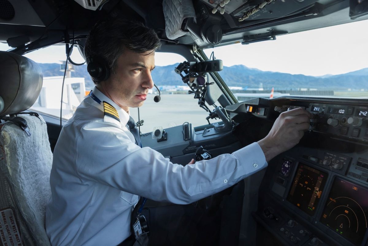 Nathan Fielder is seated in the cockpit of an airplane. He is wearing a white pilot&rsquo;s uniform and headset, and adjusting the dials on the control panel.