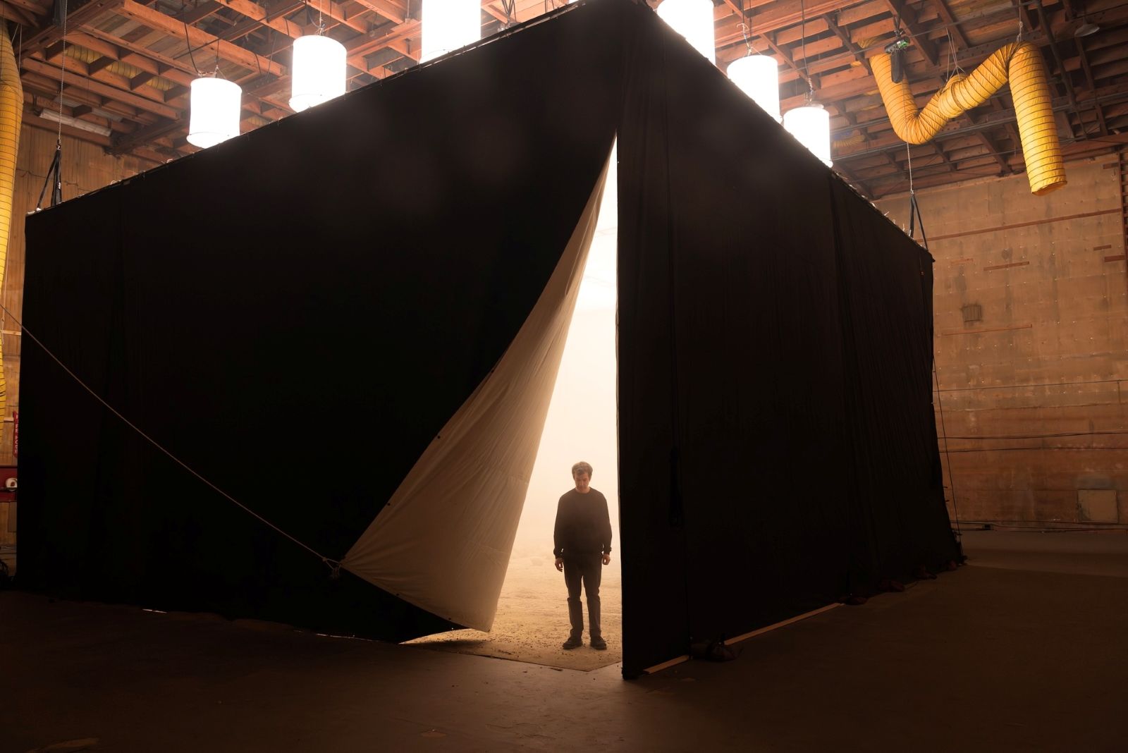 Nathan Fielder stands on a darkened soundstage in a television studio. Around him is a rectangular space whose walls are covered in dark drapery. One opens to reveal Fielder, standing in a pool of light.