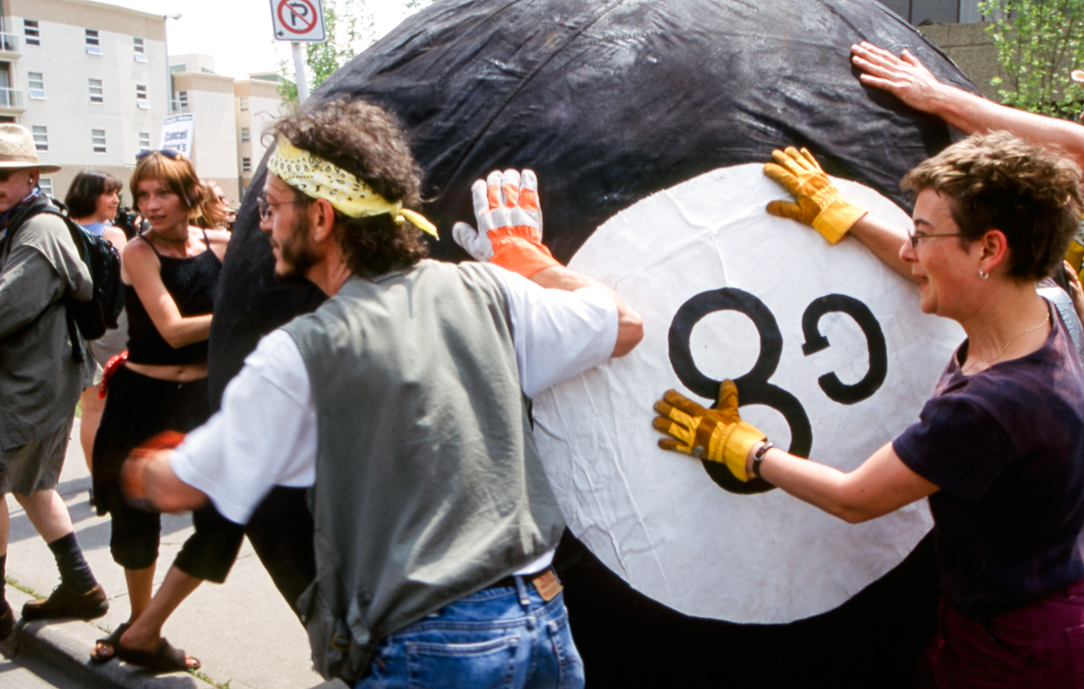 Top: protesters in a march are shot from the back. On the right, a person is wearing a black sleeveless top and a white sign is pinned to their back with the slogan, “If I can’t dance…it’s not my revolution!” Bottom: A group of people works together to roll a giant handmade “G8” eight ball down a city street.