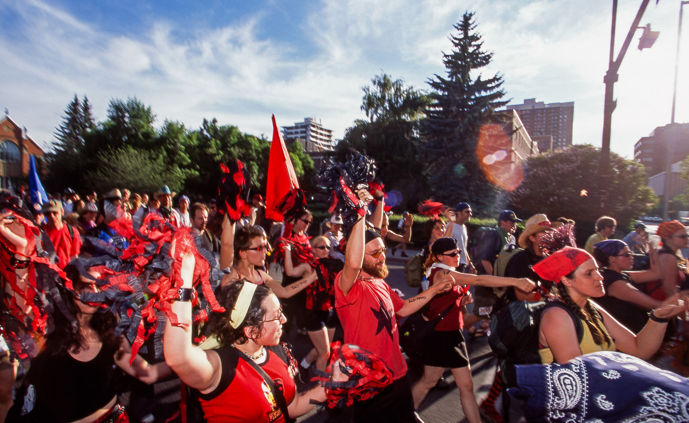 A group of protesters marches together on a sunny day. Many are wearing red and black and holding red and black flags or bunches of streamers. 